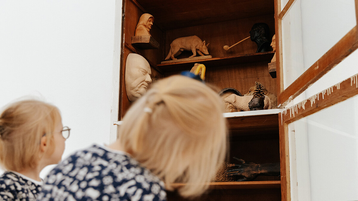 Zwei Kinder sitzen vor einem offenem Schrank mit Kunstwerken, hier Holzskulpturen, von Paloma Varga Weisz